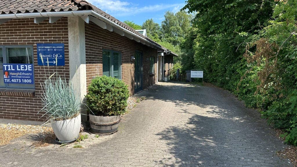 Indkørslen og den egen indgang til lokalerne på Høgevej 3, med beplantning langs bygningen og plads til parkering.
